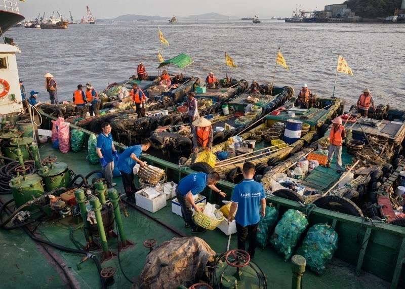 Seven small fishing boats unload plastic onto a barge for recycling.